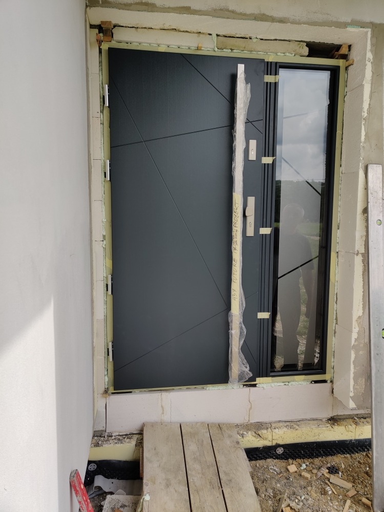 Montaż nowoczesnych drzwi wejściowych z bocznym przeszkleniem, widoczne pianka montażowa i deski zabezpieczające podłoże, budowa w trakcie wykończenia.