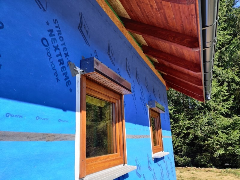 Montaż okna z roletą zewnętrzną na budynku z niebieską membraną izolacyjną, widoczne drewniane elementy konstrukcyjne dachu i rynna.