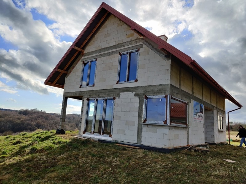 Surowy budynek jednorodzinny w trakcie montażu okien z brązowymi ramami, widoczne bloczki betonowe i czerwona dachówka, krajobraz pagórkowaty w tle.