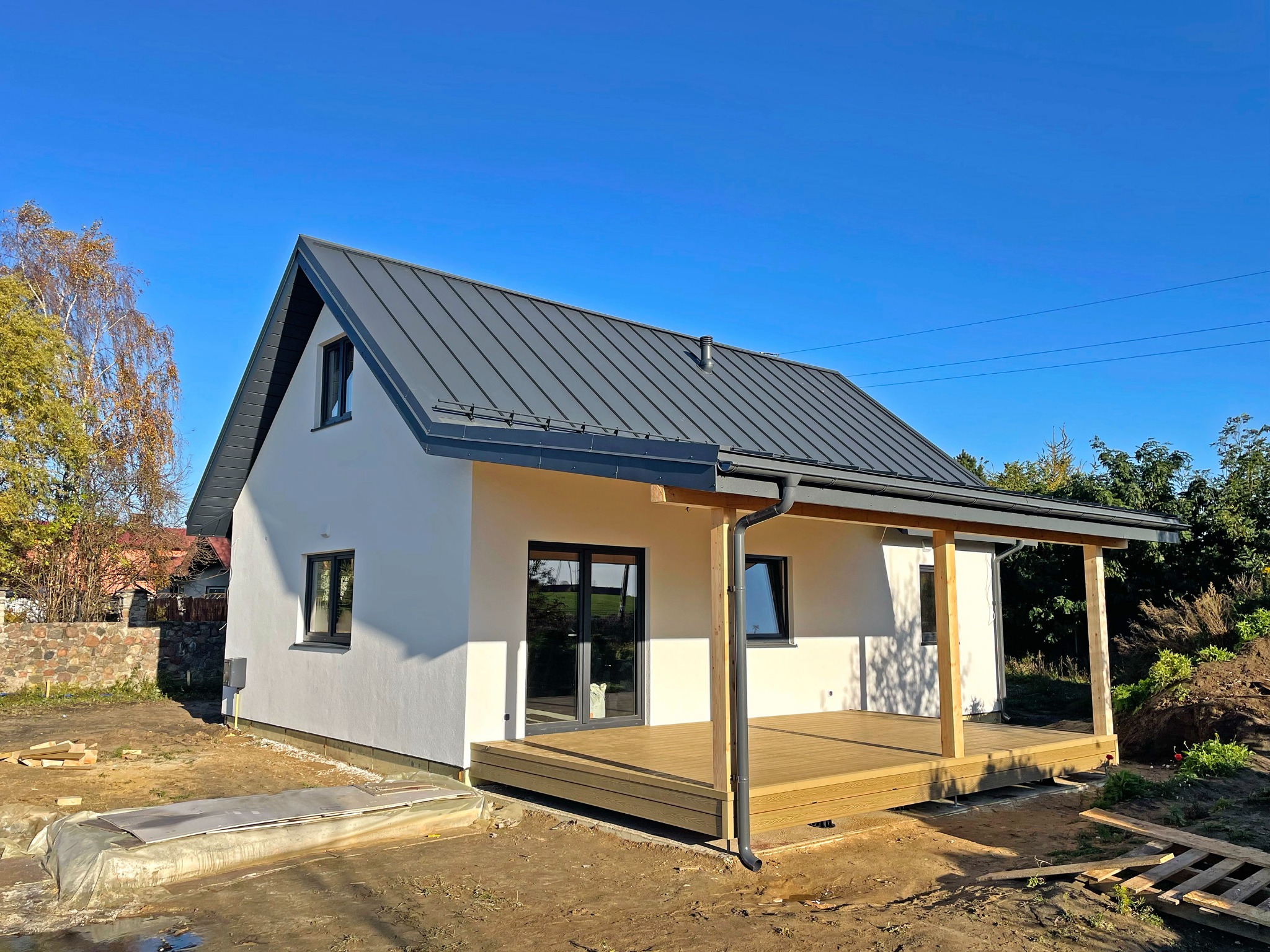 Nowoczesny dom szkieletowy z szarą blachodachówką i drewnianym tarasem w słoneczny dzień. Widoczne detale wykończenia i otoczenie w trakcie prac.