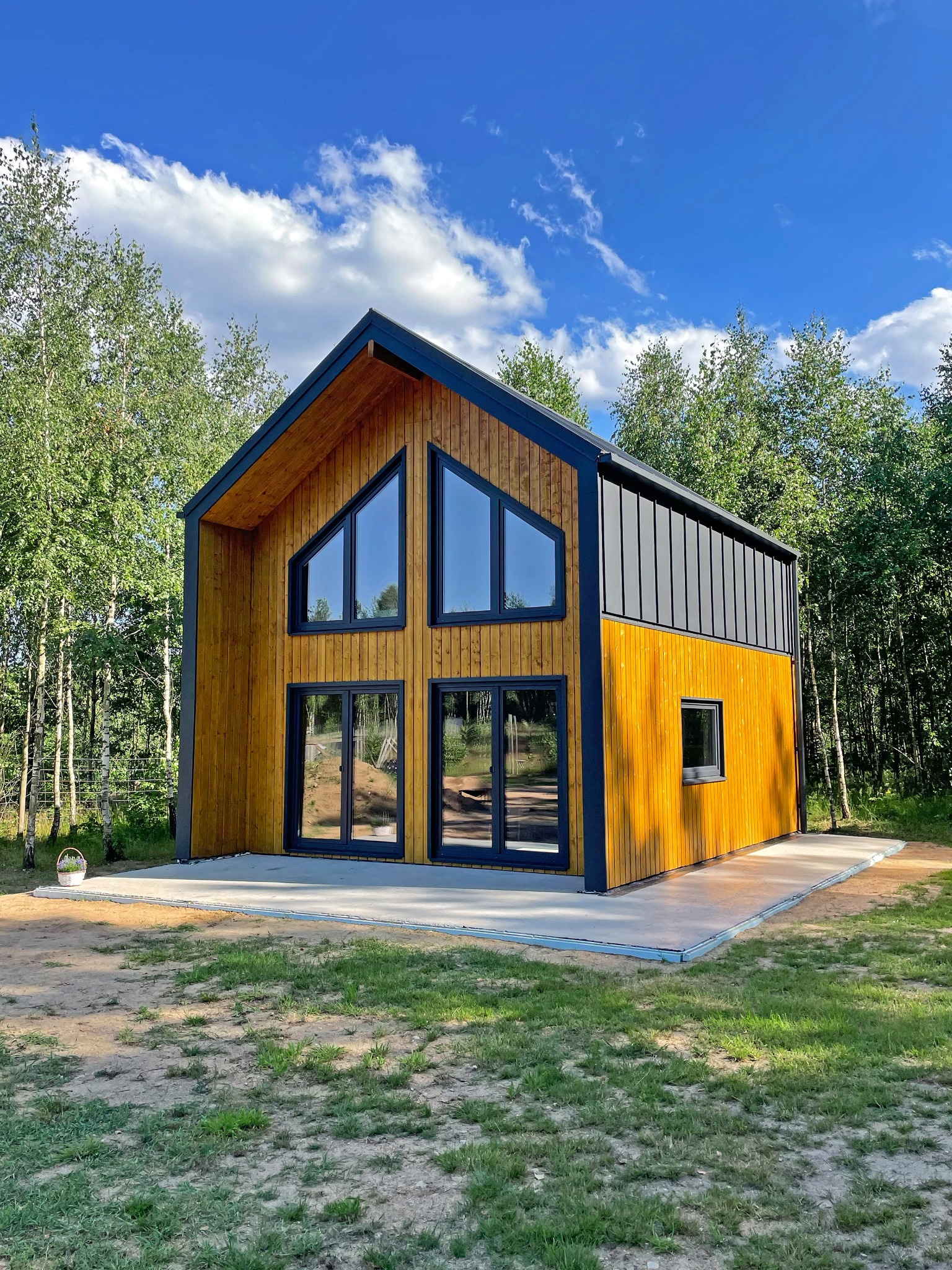 Nowoczesny dom z drewna o żółtej elewacji i czarnym dachem, otoczony zielenią drzew, prezentowany w słoneczny dzień z błękitnym niebem w tle.