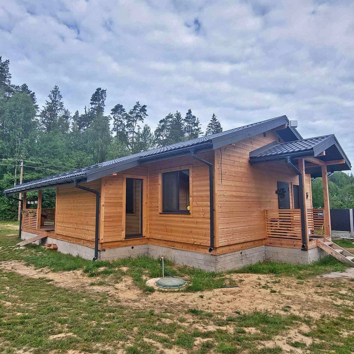 Nowoczesny, parterowy dom z drewnianą elewacją, ciemną dachówką i przeszklonymi drzwiami wejściowymi, otoczony zielenią.