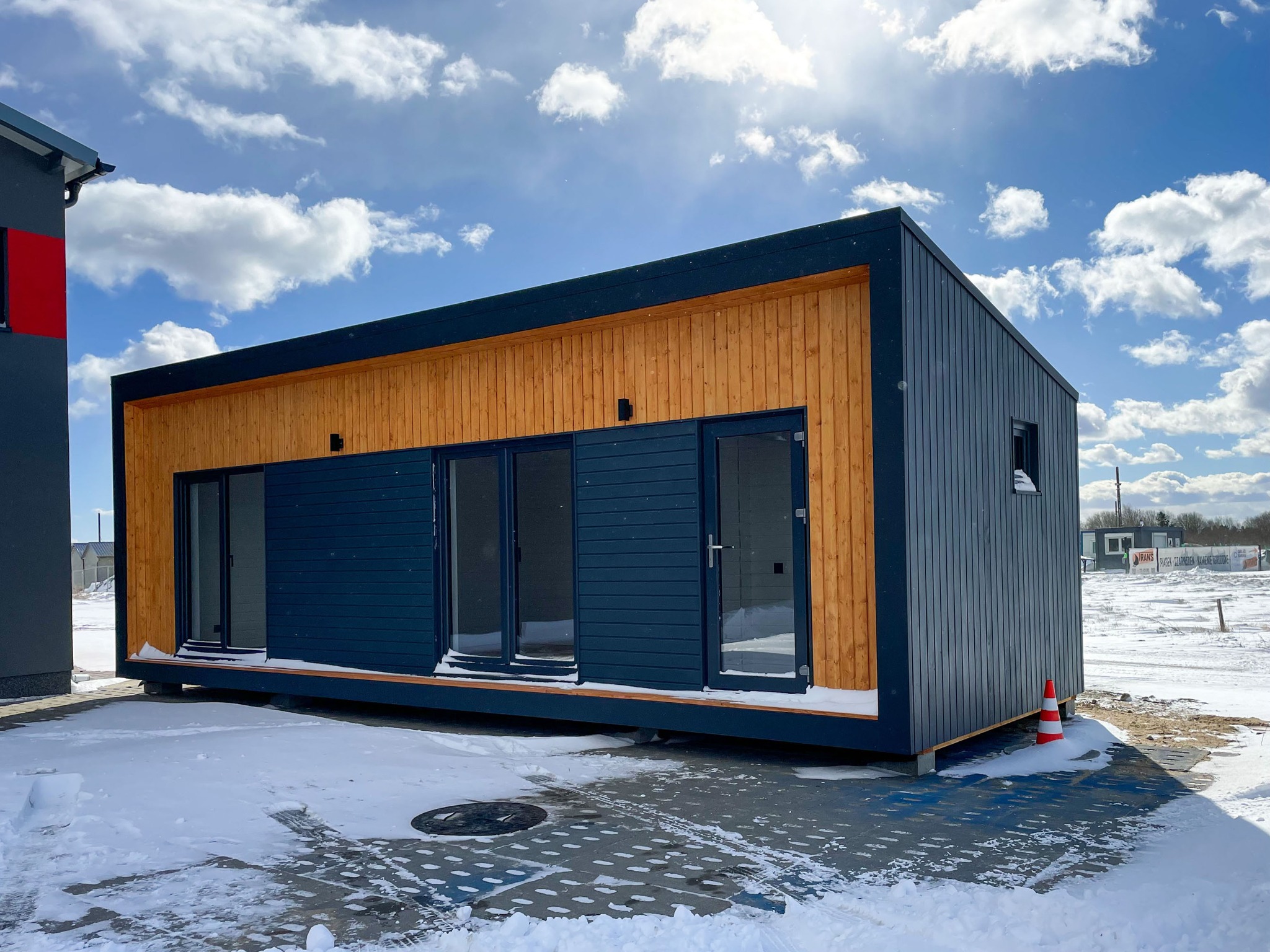 Nowoczesny dom modułowy z drewnianą elewacją i antracytowymi panelami, częściowo zasypany śniegiem, na tle błękitnego nieba z chmurami.