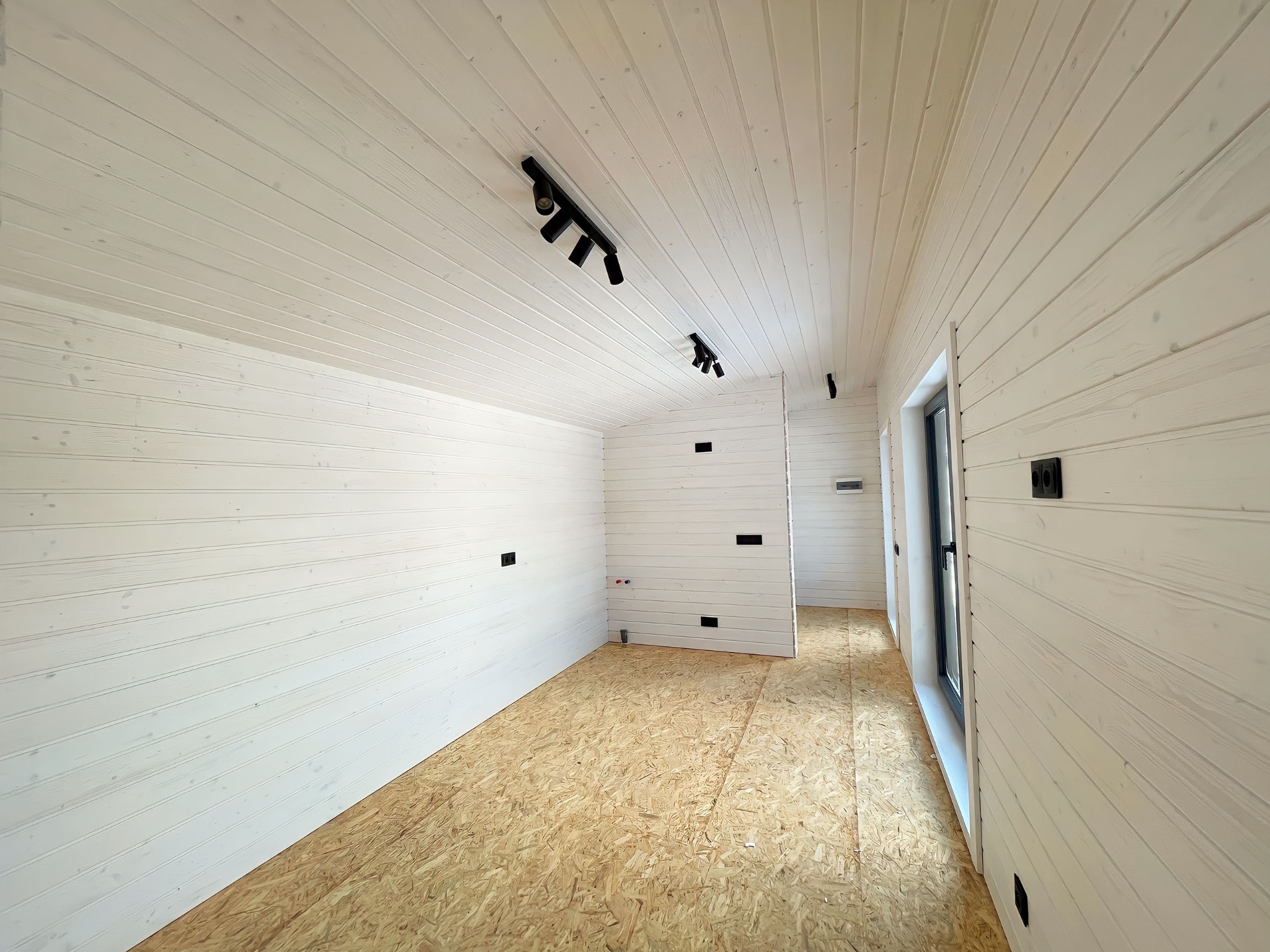 Wnętrze w trakcie wykańczania: białe, drewniane ściany z widocznymi punktami elektrycznymi, surowa podłoga z płyt OSB, czarne reflektory sufitowe i okno z czarną ramą.