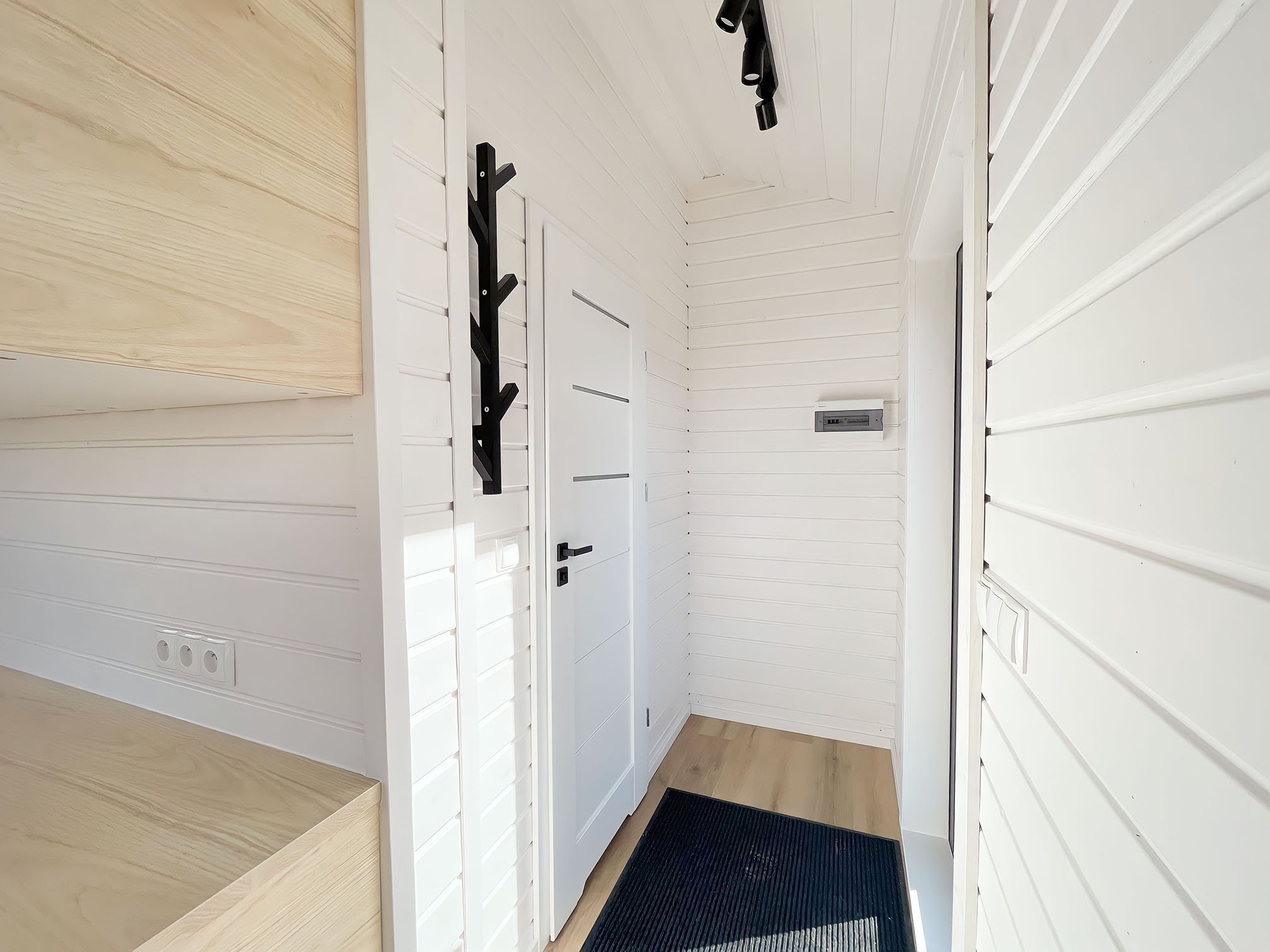 Wnętrze przedpokoju wykończonego białymi panelami boazeryjnymi, czarny wieszak na ubrania w kształcie drzewa, białe drzwi wewnętrzne z trzema poziomymi frezowaniami, czarna wycieraczka, jasne...