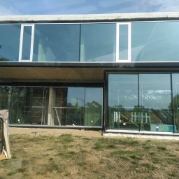 Okna Cichosza - Nowoczesny budynek z dużymi, panoramicznymi oknami aluminiowymi, widoczne rusztowania i przygotowane szyby. Prace montażowe w toku, ziemisty teren wokół.
