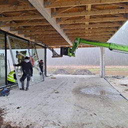 Okna Cichosza - Montaż panoramicznego okna z użyciem przyssawki i dźwigu na budowie domu. Pracownicy w kamizelkach odblaskowych, drewniana konstrukcja dachu.