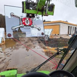 Okna Cichosza - Montaż dużej tafli szklanej za pomocą przyssawki na dźwigu, w tle budynek w stanie surowym otwartym i teren budowy po deszczu. Widok z kabiny operatora.