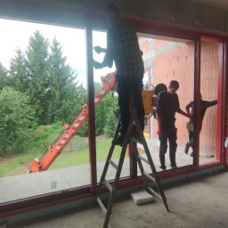 Okna Cichosza - Montaż okna panoramicznego z czerwoną ramą w surowym wnętrzu. Na zewnątrz widoczny dźwig i zieleń. Pracownicy na drabinie i wewnątrz pomieszczenia.