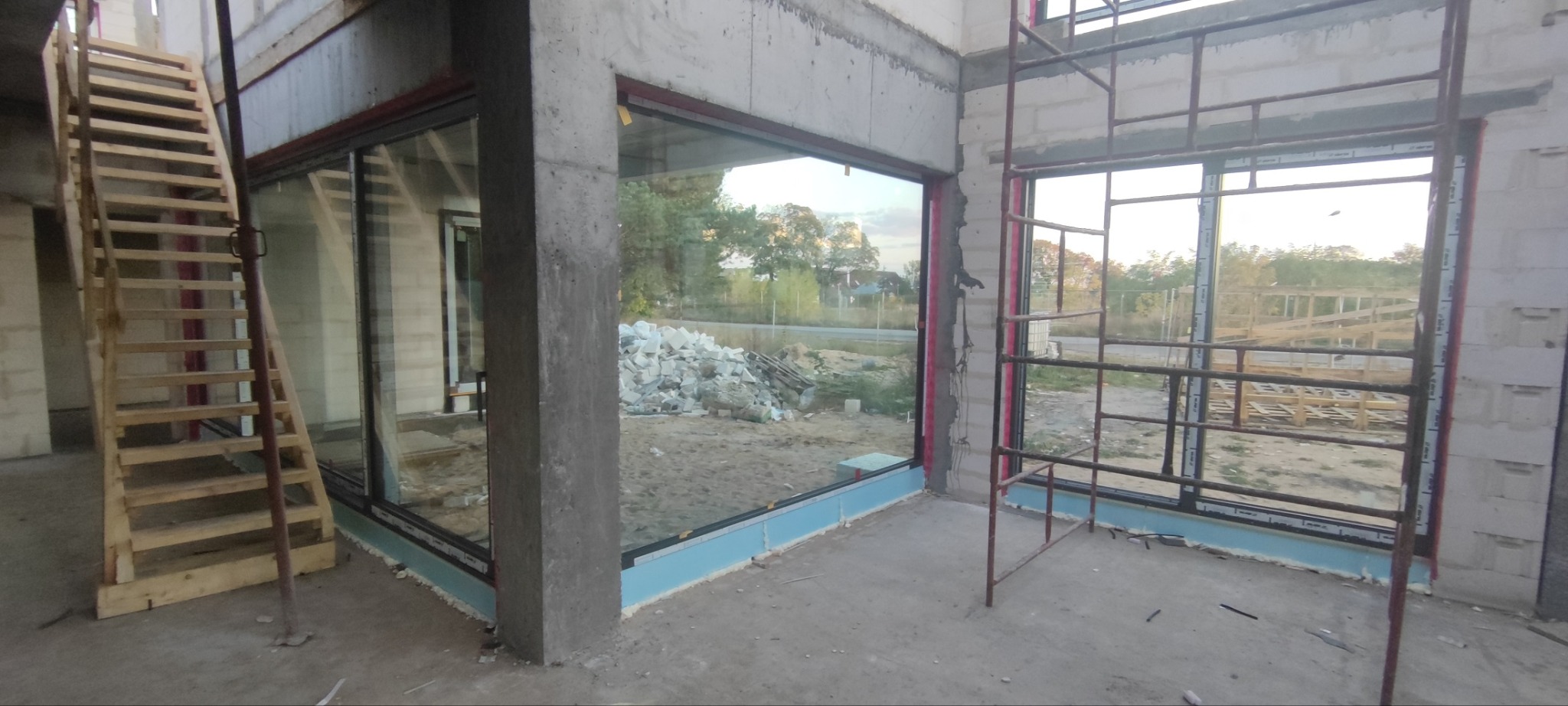 Montaż okien PCV w surowym stanie budynku, widoczne rusztowania, drewniane schody i materiały budowlane na zewnątrz.