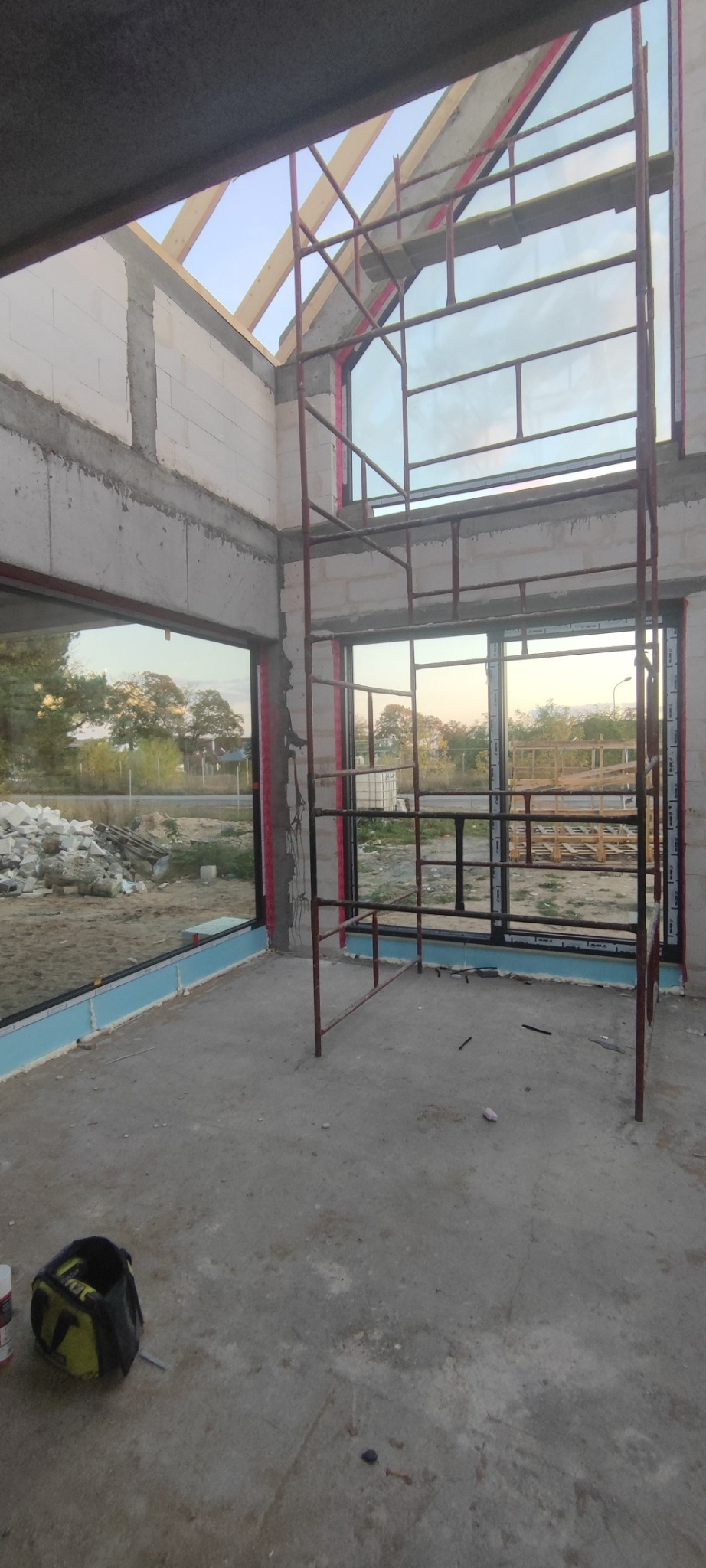 Wnętrze surowego budynku w trakcie montażu dużych okien z widokiem na zewnątrz, rusztowanie w środku pomieszczenia, torba narzędziowa na podłodze.