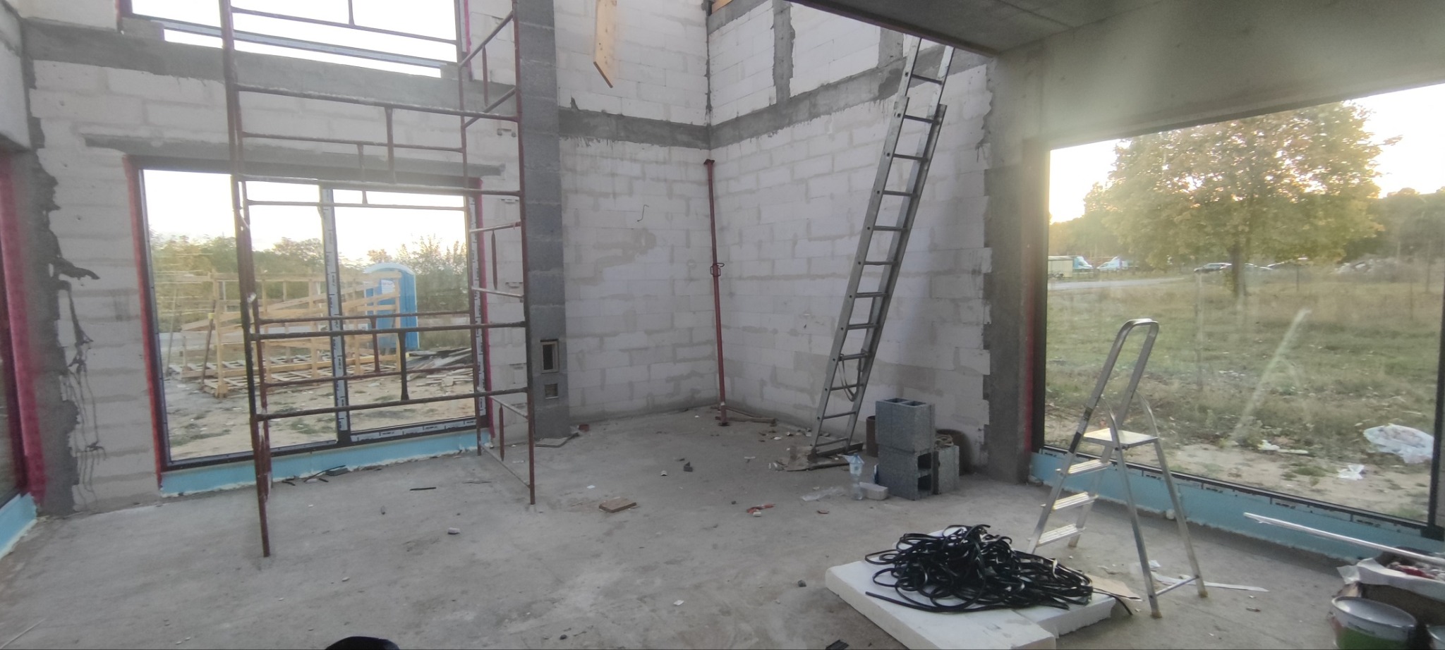 Wnętrze budynku w trakcie budowy, widoczne surowe ściany z betonu komórkowego, częściowo zamontowane ramy okienne z widokiem na zewnątrz, metalowe rusztowania, drabiny, oraz materiały budowlane...