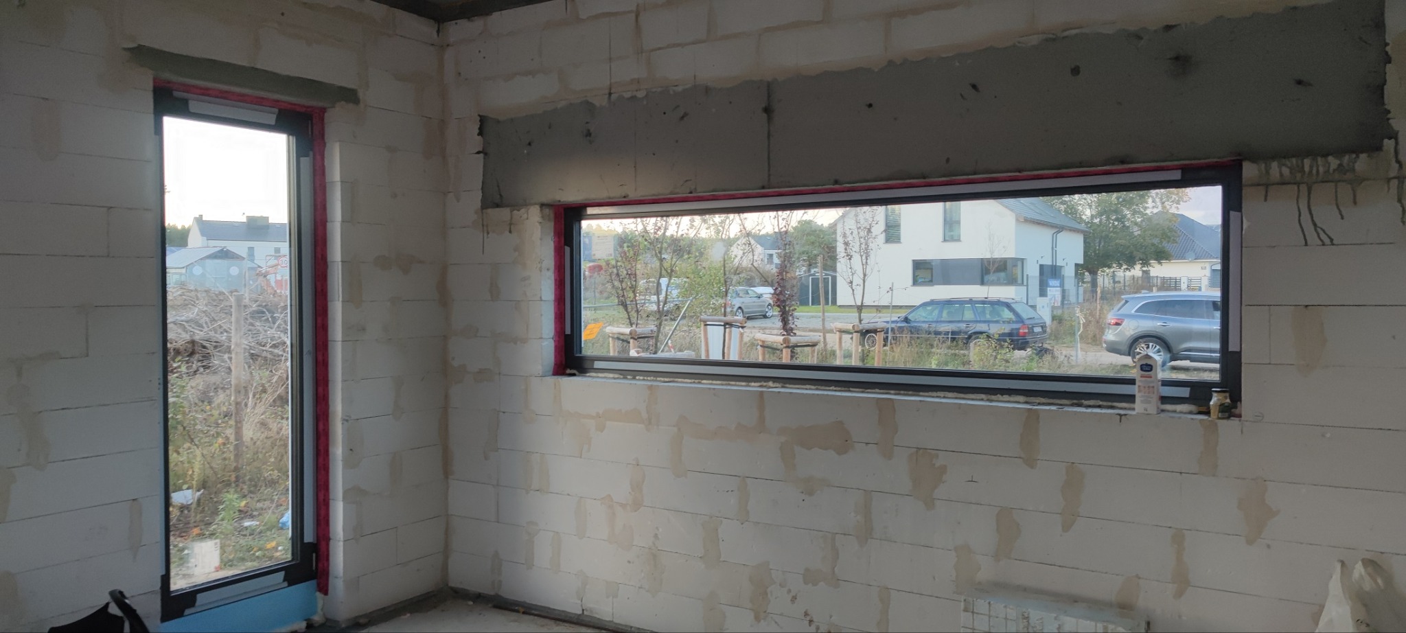 Surowy stan: dwa okna z czarnymi ramami w ścianie z białych bloczków, widok na zewnątrz z samochodami i domami, zaprawa pod oknem, butelka i słoik na parapecie.