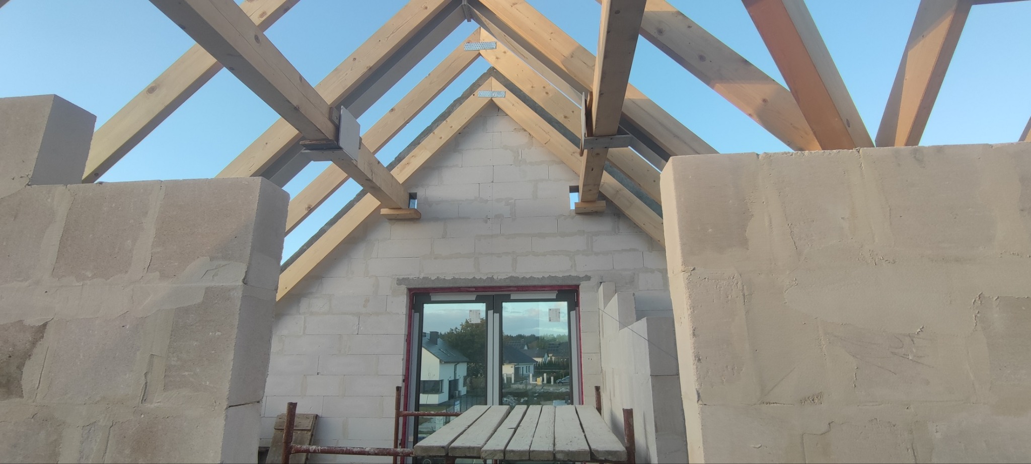 Surowy stan budynku w trakcie budowy: ściany z betonu komórkowego, drewniana konstrukcja dachu, okno z widokiem na zabudowania w tle.