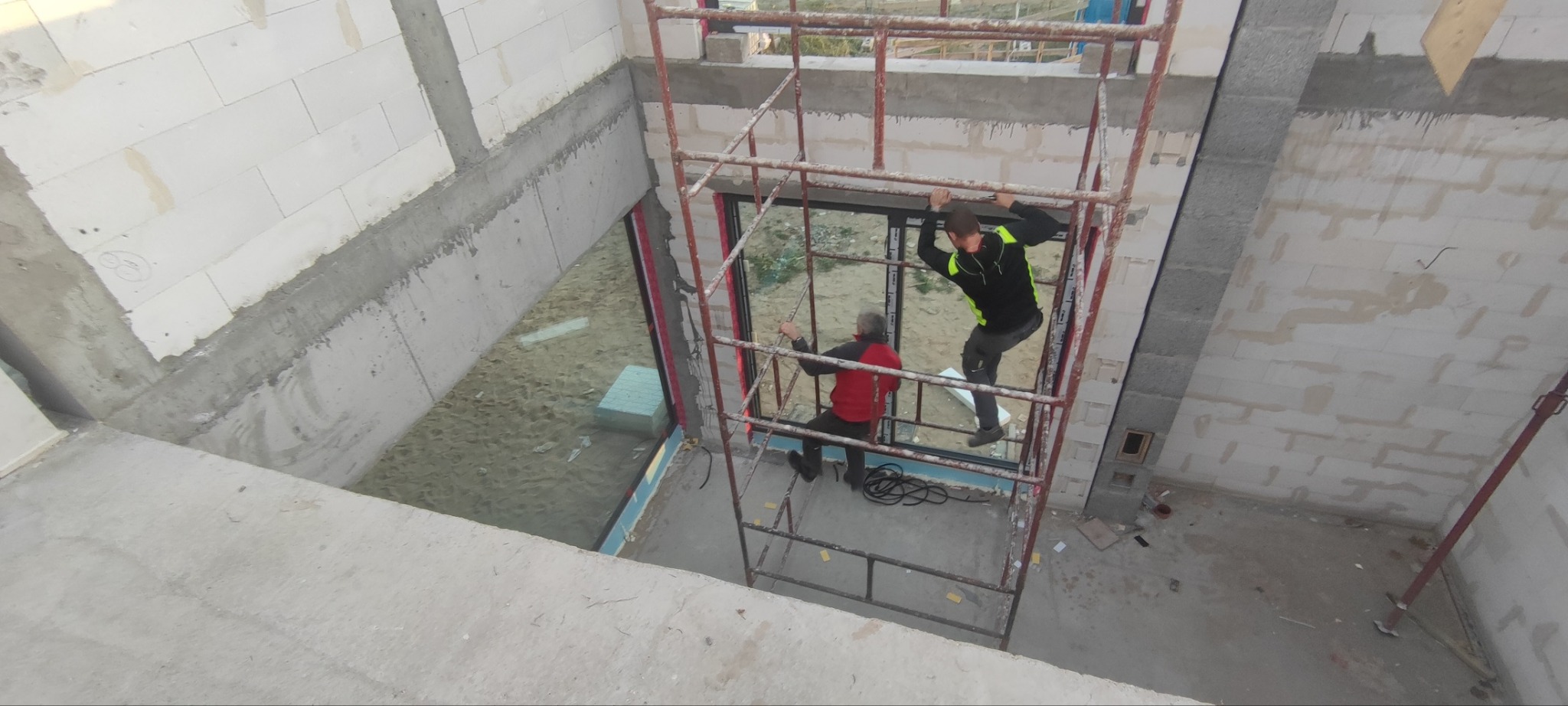 Montaż okna podczas budowy domu: dwóch monterów na rusztowaniu, widok z góry na surowy stan budynku z pustaków.