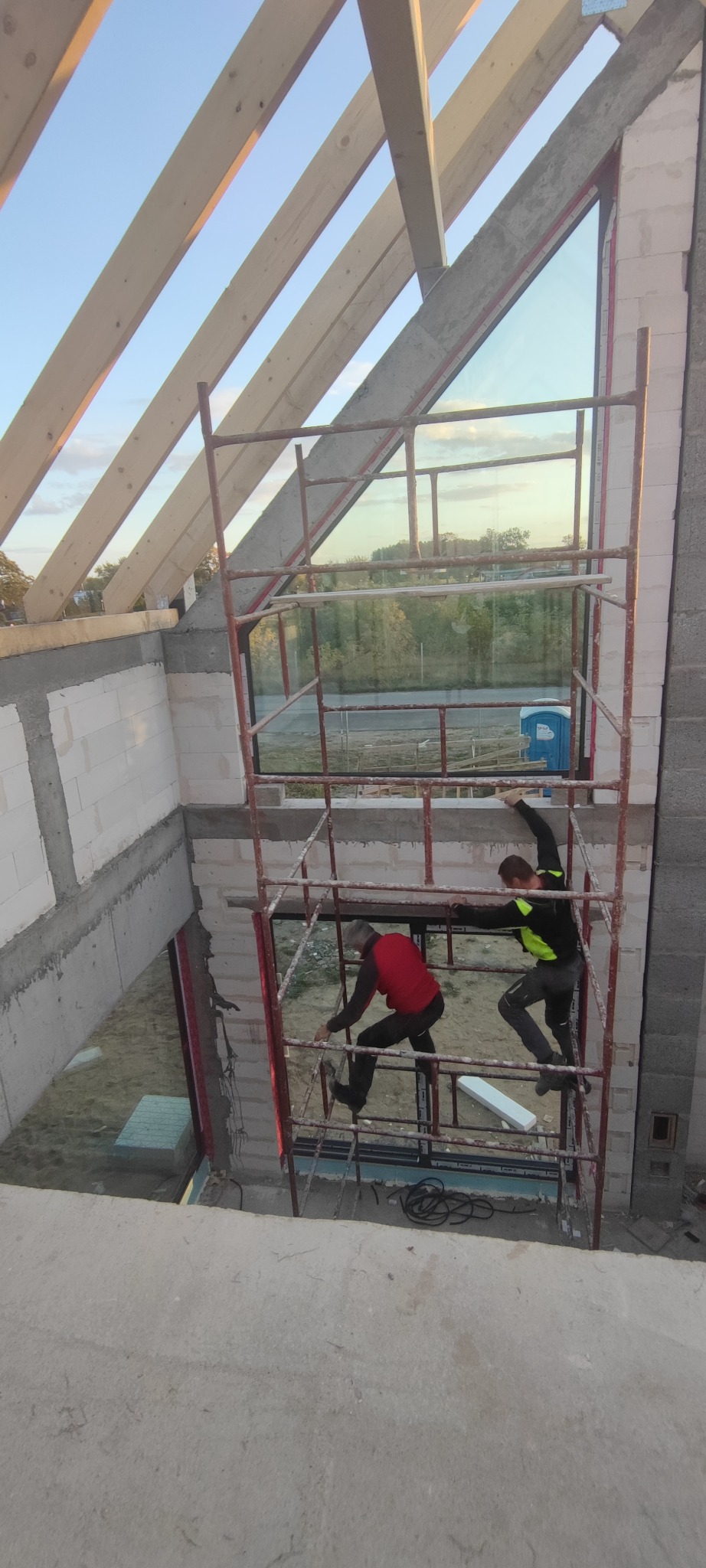 Dwóch monterów na rusztowaniu instaluje okno w surowym stanie budynku, widoczne drewniane elementy konstrukcji dachu i fragment otoczenia za oknem.