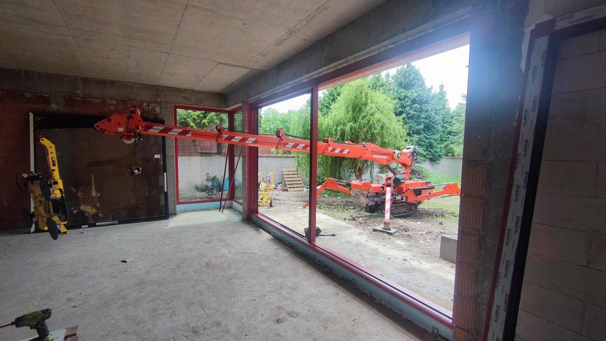 Montaż okna za pomocą miniżurawia w surowym wnętrzu. Widoczne narzędzia, ramy okienne i zielone otoczenie zewnętrzne. Poznań k/ Pestki.