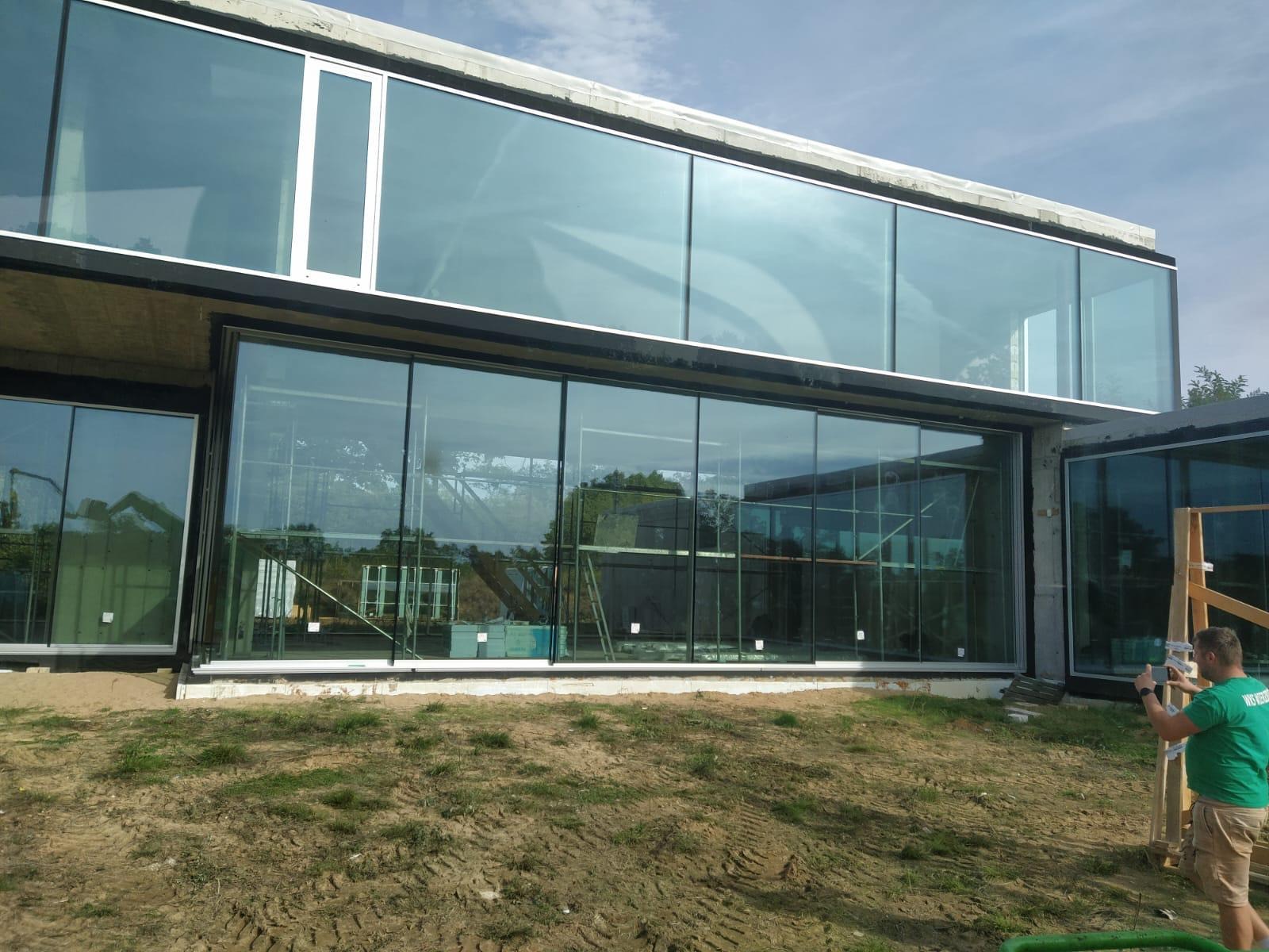 Nowoczesny budynek z dużymi przeszkleniami, widoczne odbicia otoczenia, częściowo wykończony teren wokół budynku. Na pierwszym planie ziemia.