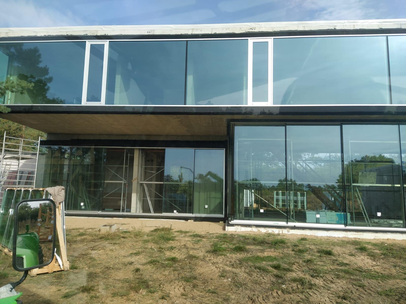 Nowoczesny budynek z dużymi, panoramicznymi oknami aluminiowymi, widoczne rusztowania i przygotowane szyby. Prace montażowe w toku, ziemisty teren wokół.