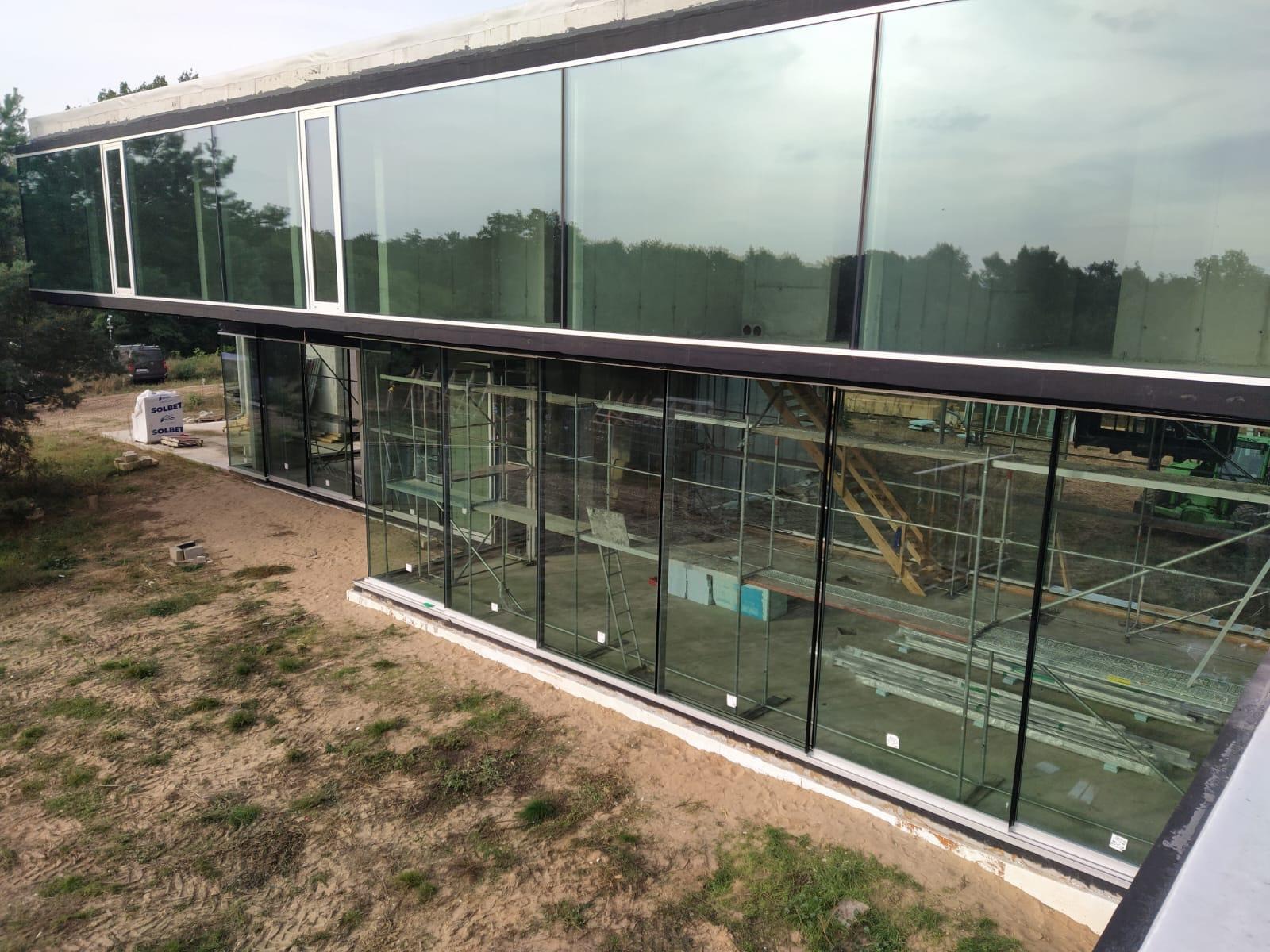 Montaż panoramicznych okien w nowoczesnym budynku. Widoczne rusztowania i teren budowy. Duże przeszklenia odbijają otoczenie.