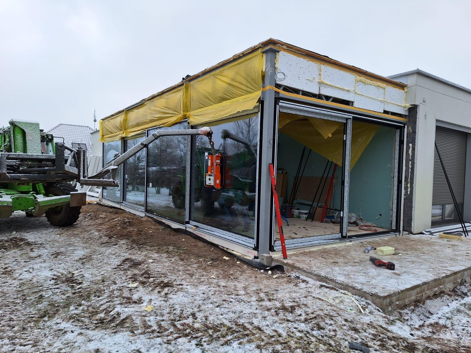 Montaż panoramicznych okien przesuwnych w nowym budynku z użyciem dźwigu. Widoczny poziomica, narzędzia i ośnieżone otoczenie.