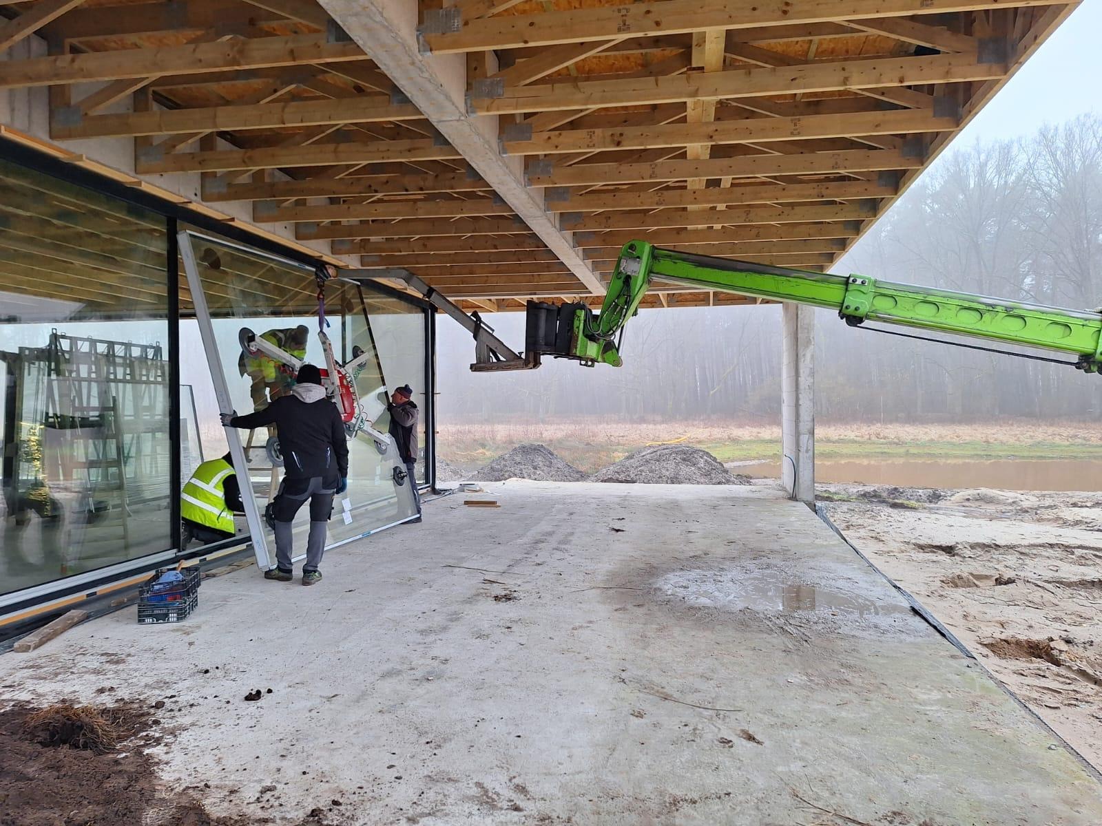 Montaż panoramicznego okna z użyciem przyssawki i dźwigu na budowie domu. Pracownicy w kamizelkach odblaskowych, drewniana konstrukcja dachu.