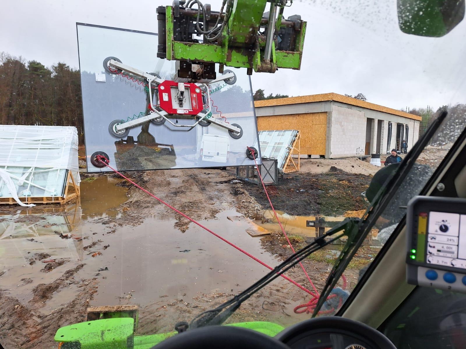 Montaż dużej tafli szklanej za pomocą przyssawki na dźwigu, w tle budynek w stanie surowym otwartym i teren budowy po deszczu. Widok z kabiny operatora.