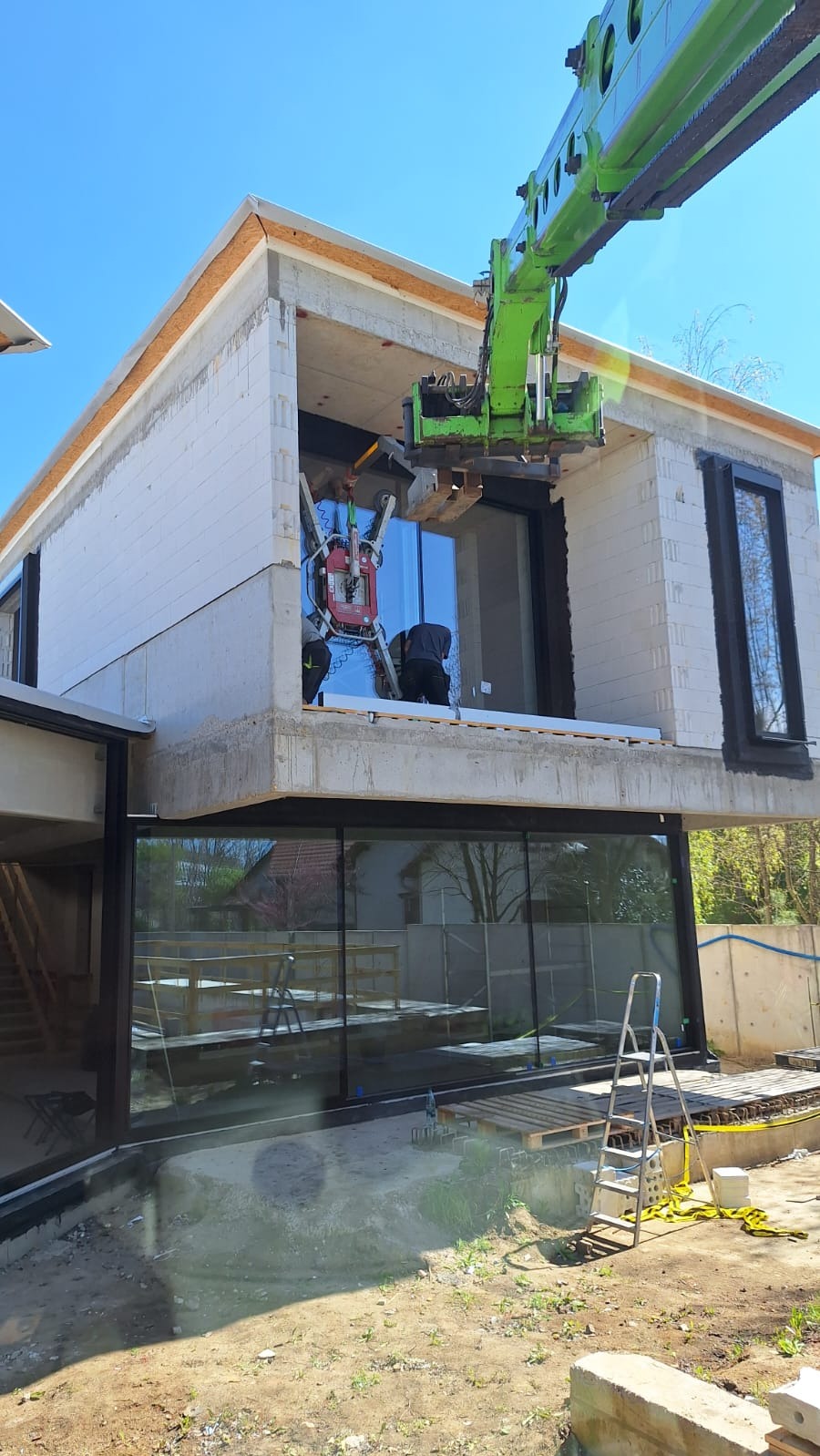 Montaż okna za pomocą dźwigu na placu budowy domu. Widoczne narzędzia i pracownicy w trakcie instalacji. Duże przeszklenia na dwóch kondygnacjach.