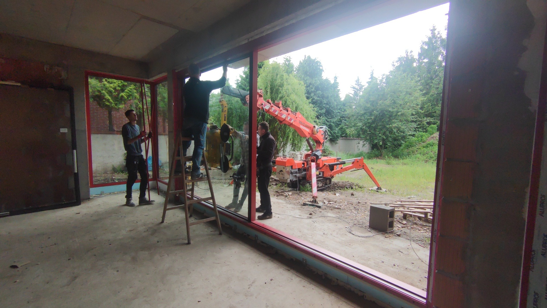 Montaż okien z użyciem mini żurawia na budowie. Widoczni trzej monterzy i częściowo zamontowane okno z czerwoną ramą. Na zewnątrz zieleń i sprzęt budowlany.