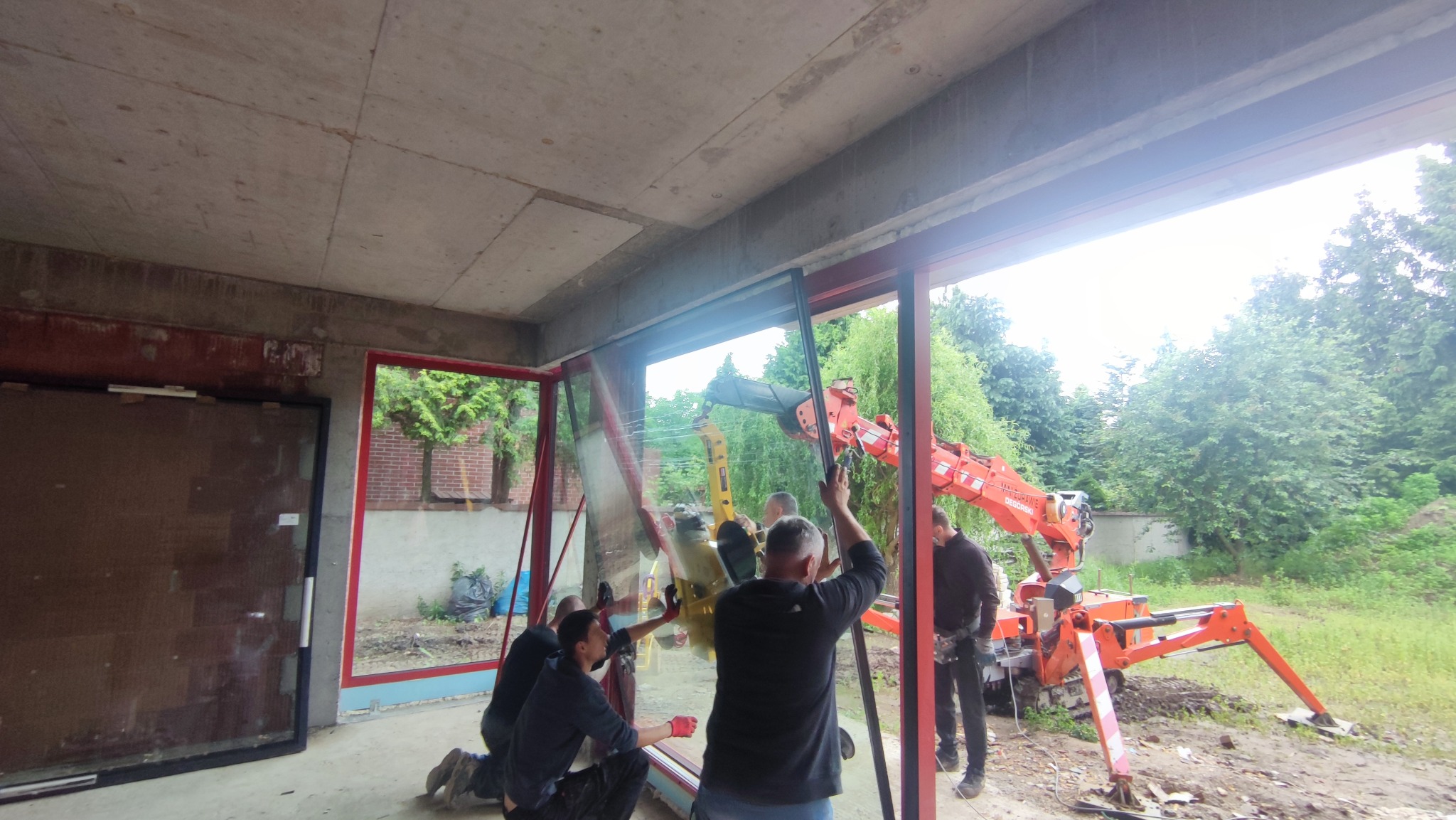 Montaż okna przez ekipę w Poznaniu, z użyciem dźwigu. Widoczne surowe wnętrze i zieleń na zewnątrz. Praca w toku, okno w trakcie instalacji.