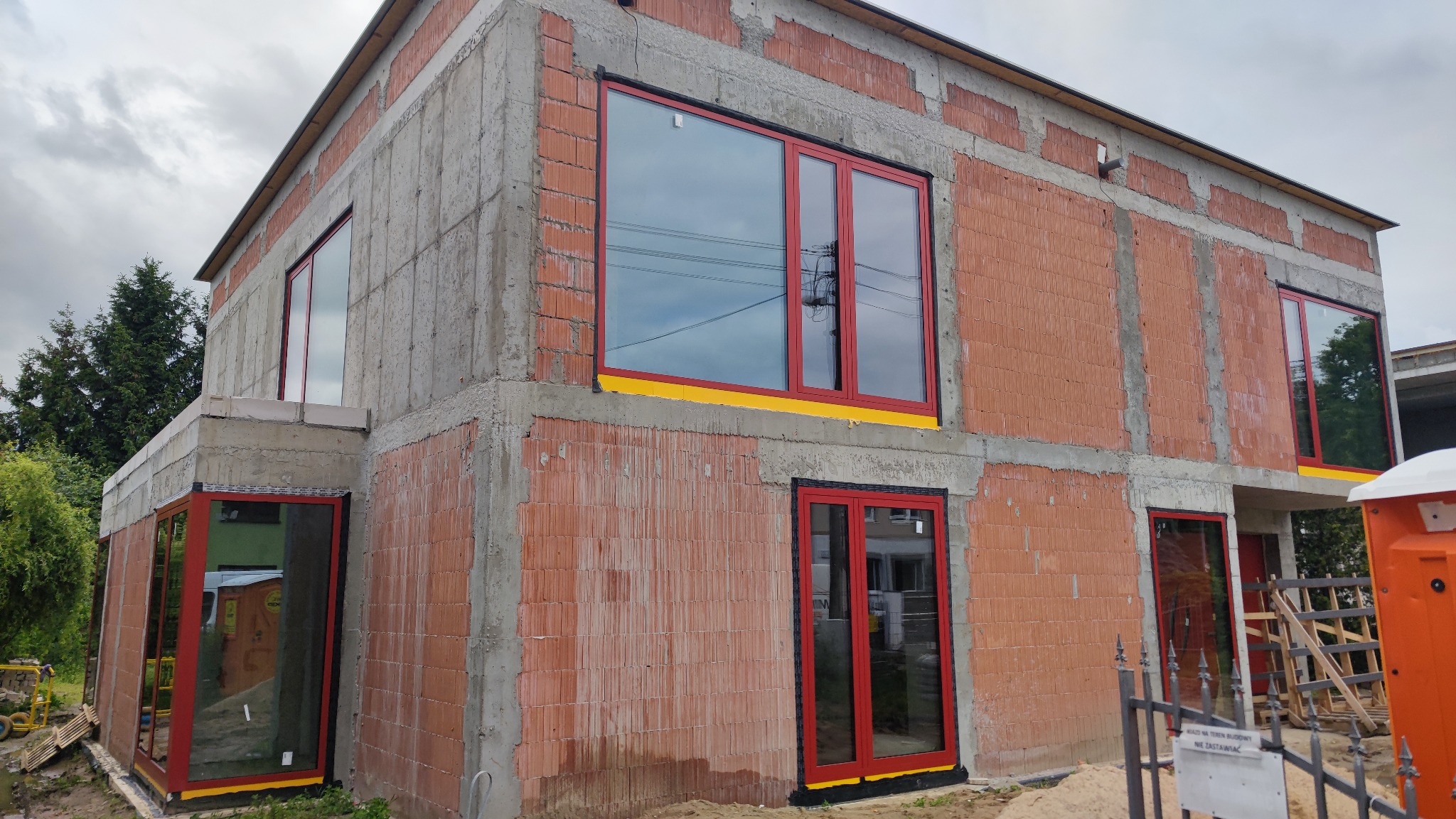 Surowy budynek w trakcie budowy z zamontowanymi oknami z czerwonymi ramami. Widoczne pustaki i betonowe wzmocnienia. Na dole żółta folia zabezpieczająca.