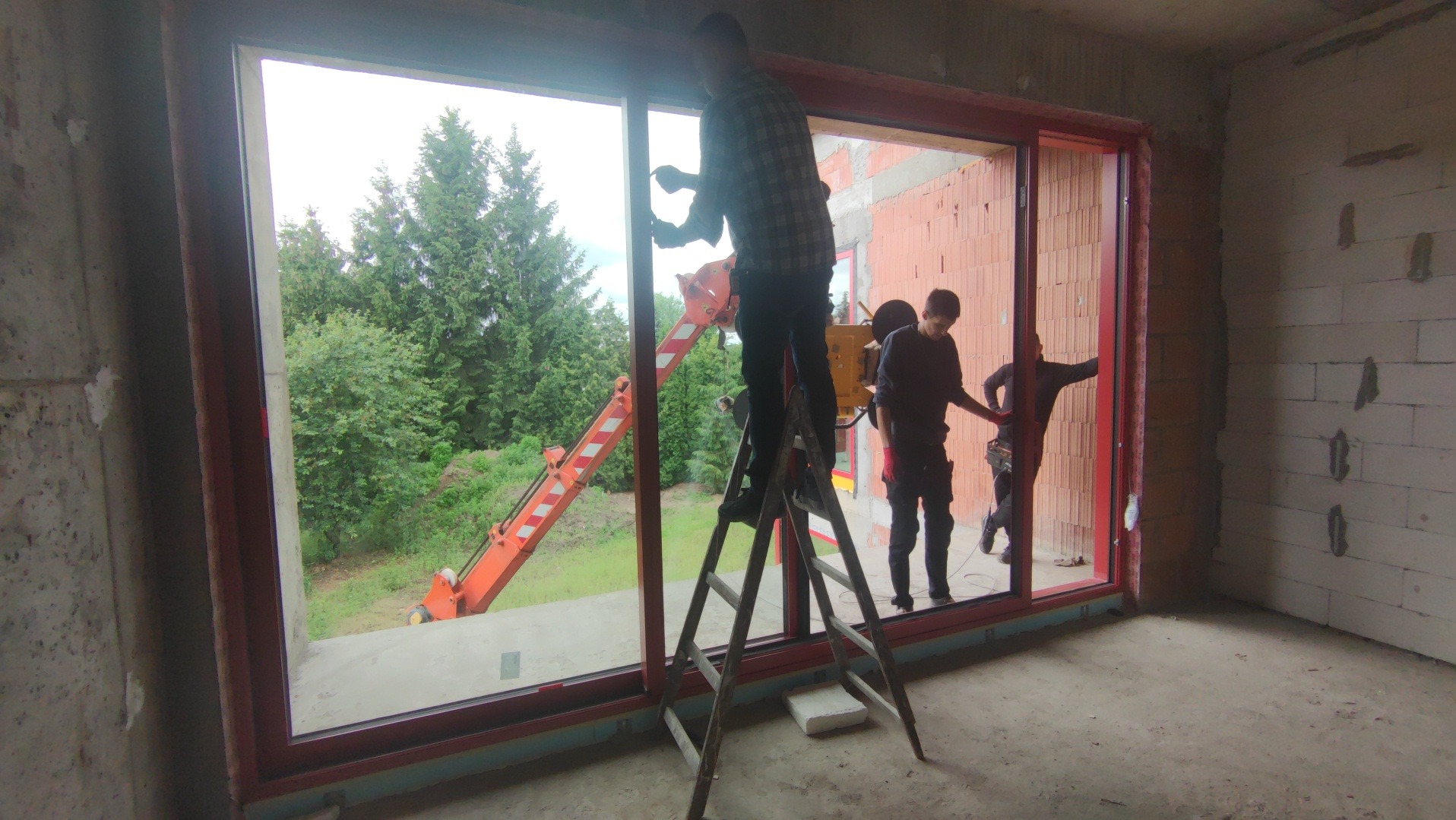 Montaż okna panoramicznego z czerwoną ramą w surowym wnętrzu. Na zewnątrz widoczny dźwig i zieleń. Pracownicy na drabinie i wewnątrz pomieszczenia.