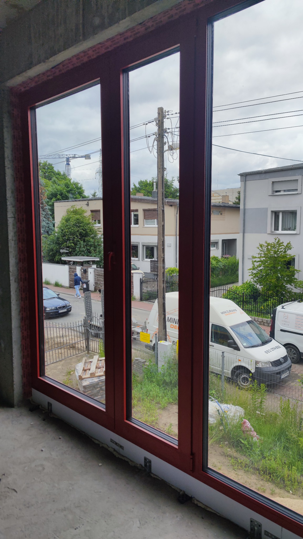 Zamontowane okno z trzema szybami w czerwonej ramie, widok z wnętrza na budynki mieszkalne i samochody na ulicy. Widoczne elementy montażowe.