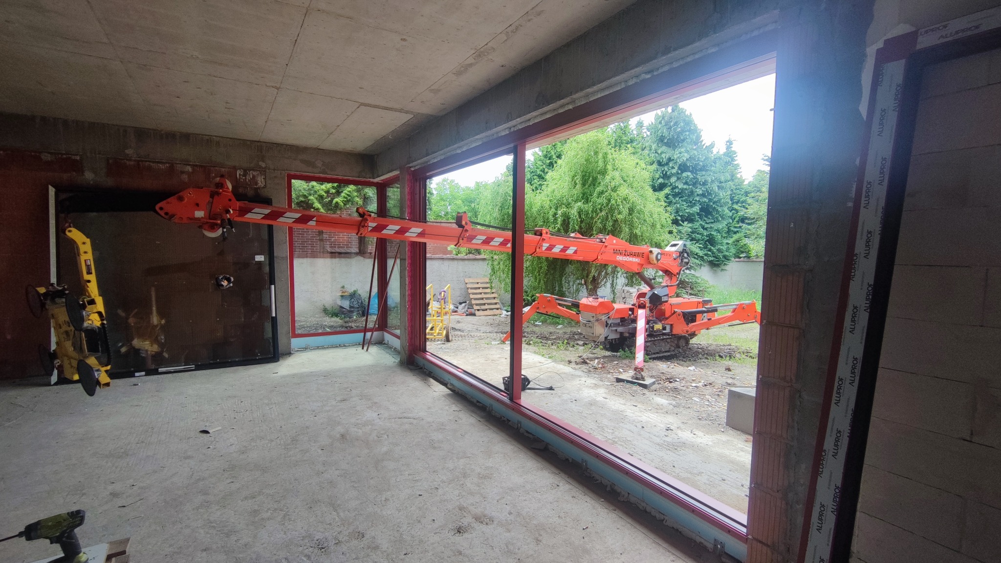 Montaż okna z użyciem pomarańczowego mini-żurawia w surowym stanie budynku. Widoczne narzędzia pracy i ramy okienne z napisem ALUPROF.