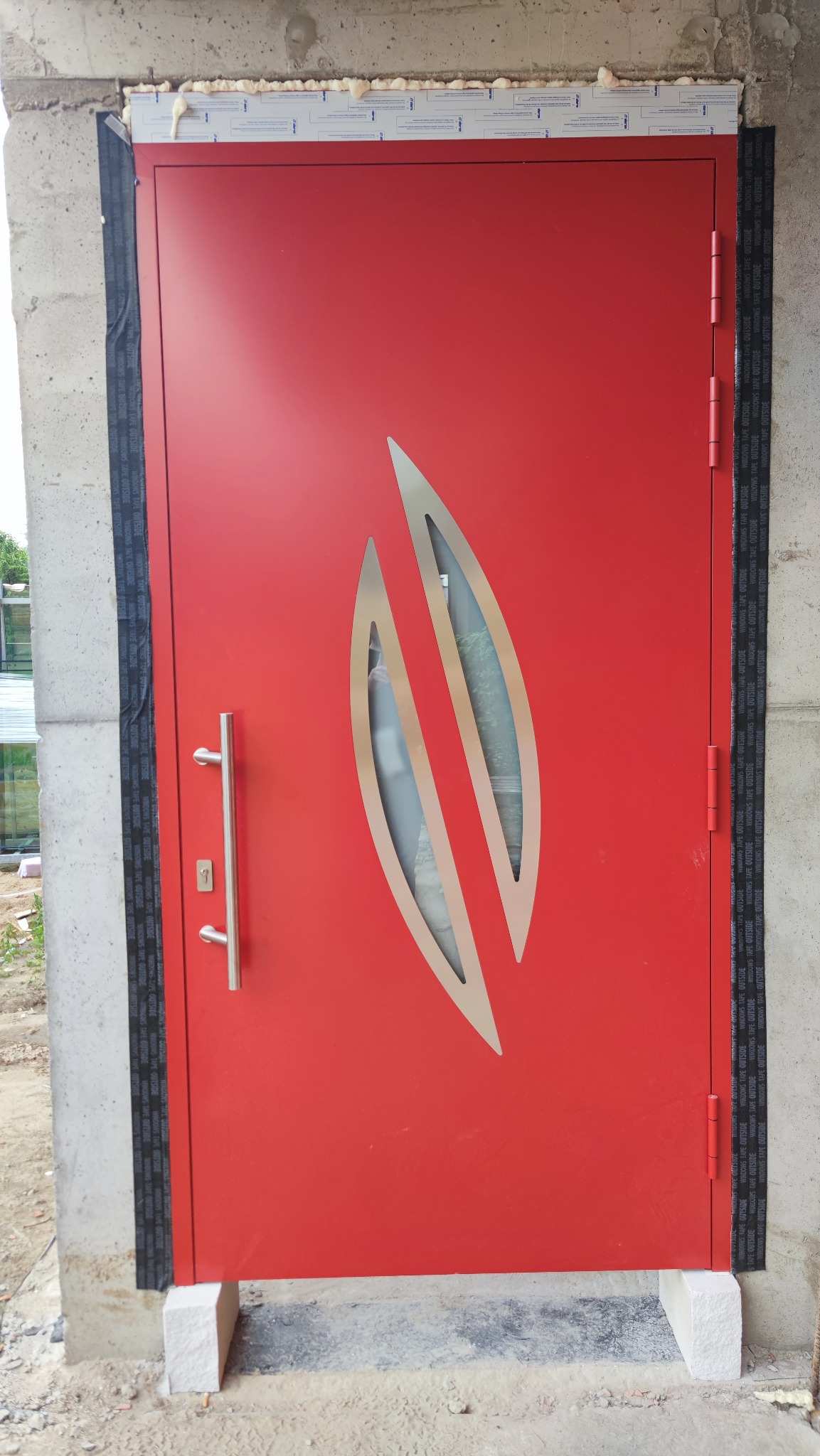 Czerwone drzwi antywłamaniowe z dekoracyjnym, podłużnym przeszkleniem, zamontowane w surowym otworze, zabezpieczone taśmą uszczelniającą. Widoczne detale montażowe.