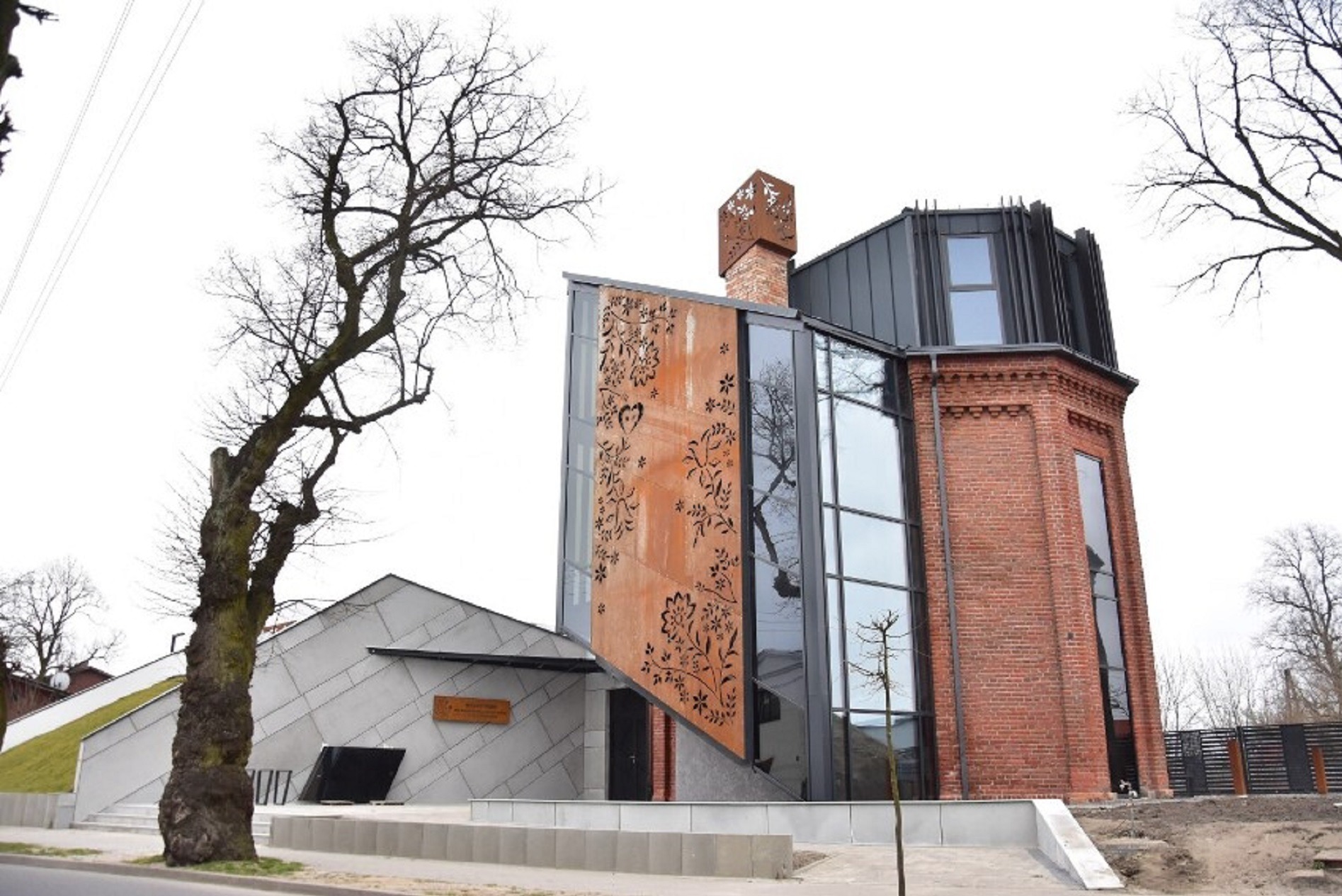 Nowoczesny budynek z elementami cegły i cortenu z wycinanym wzorem, duże okna, minimalistyczna bryła, drzewo bez liści na pierwszym planie.