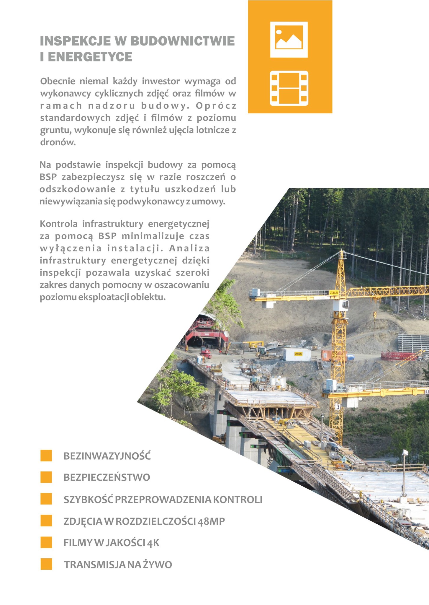 Zdjęcie lotnicze placu budowy z widocznymi dźwigami i konstrukcją mostu w trakcie realizacji, otoczonego lasem, wykonane w jasny dzień.