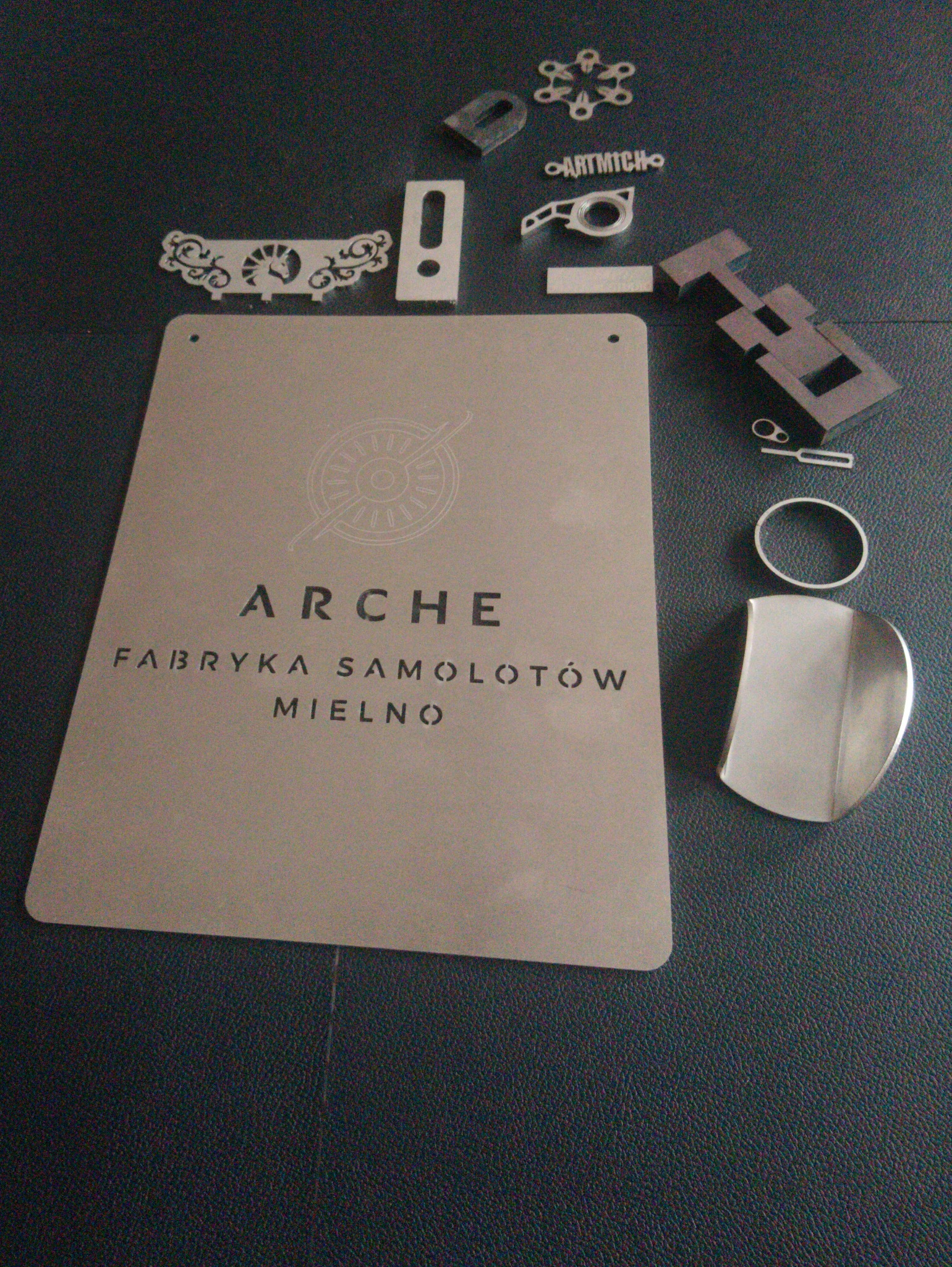 Metalowa tabliczka z napisem 'ARCHE Fabryka Samolotów Mielno' oraz wycięte laserowo elementy metalowe o różnych kształtach na ciemnym tle.
