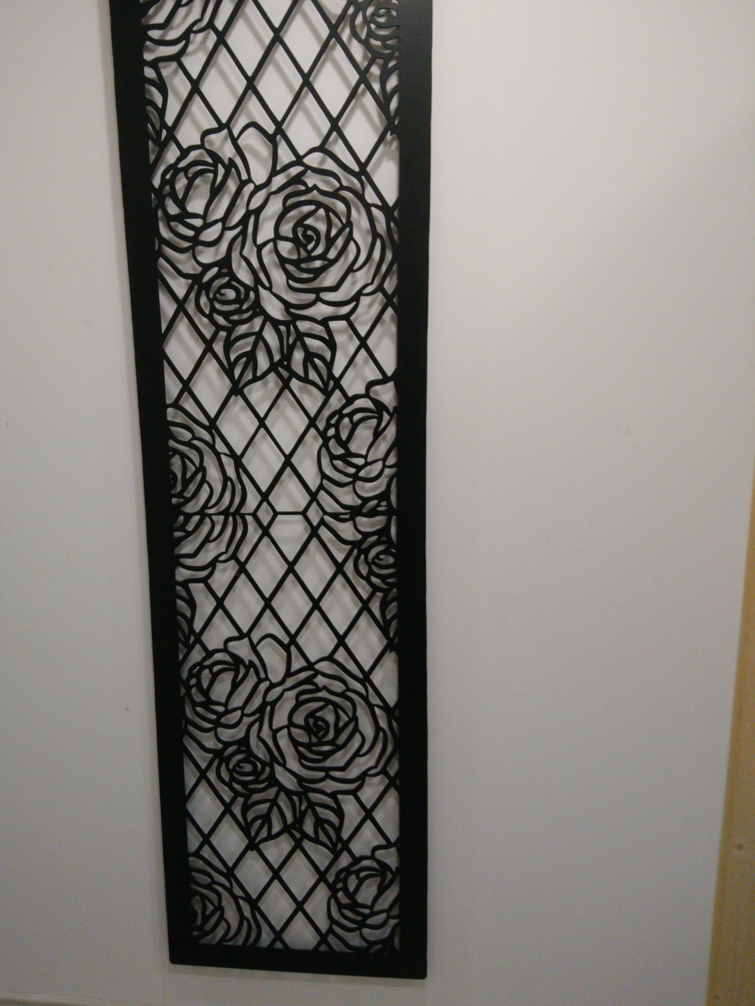 Dekoracyjny panel metalowy z motywem róż, czarny, ażurowy, przymocowany do białej ściany. Precyzyjne cięcie CNC tworzy wzór kraty i kwiatów.