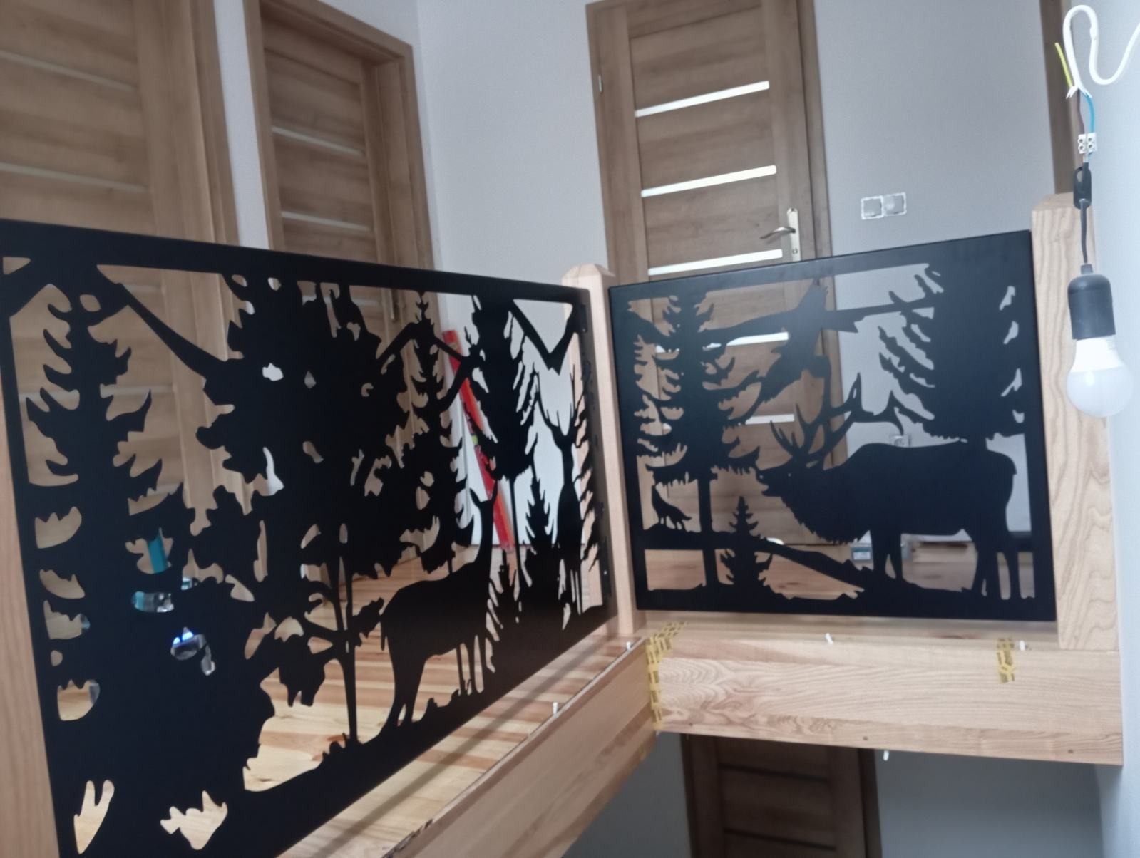 Metalowa balustrada schodowa z motywem leśnym: sylwetki drzew, jelenia i wilka, czarny metal na jasnym drewnie, widok z bliska na tle drzwi.