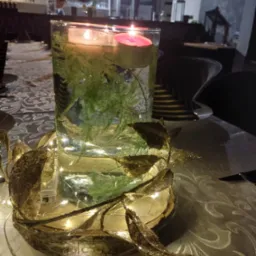 Dekoracja stołu: szklany wazon z zanurzonymi gałązkami i pływającymi świeczkami, otoczony złotą girlandą z lampkami na drewnianej podstawie, na wzorzystym obrusie.