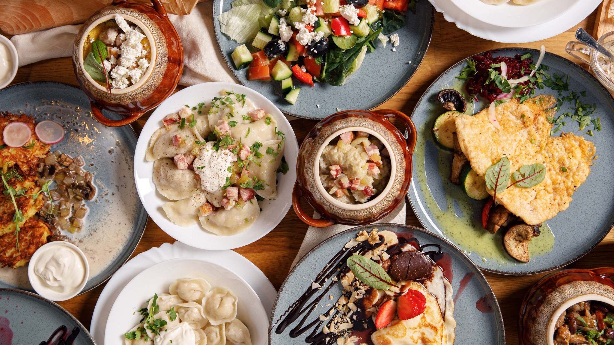 Widok z góry na różnorodne dania kuchni polskiej i europejskiej, w tym pierogi, placki ziemniaczane, sałatka grecka i kotlet schabowy, podane na talerzach i w glinianych naczyniach na drewnianym...