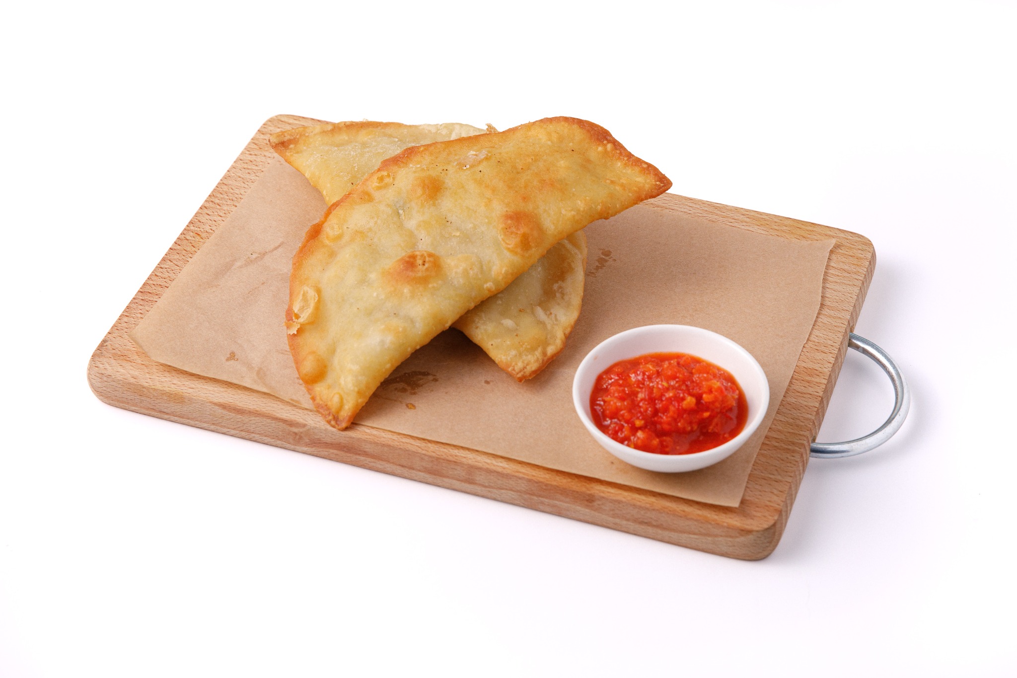 Dwa smażone pierogi na drewnianej desce z papierem pergaminowym, obok mała miseczka z czerwonym sosem pomidorowym.