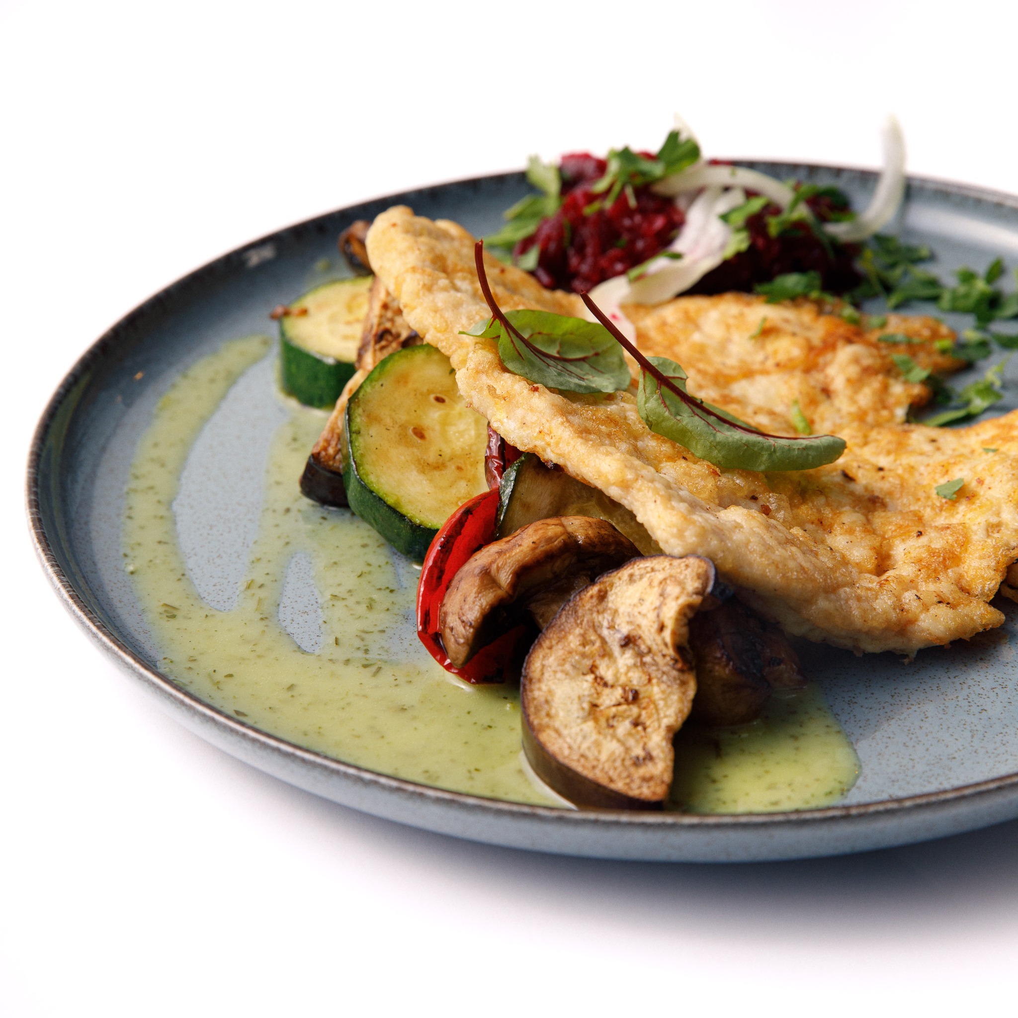Delikatnie panierowana ryba z grillowanymi warzywami (bakłażan, cukinia, papryka) polana zielonym sosem i udekorowana sałatką z buraka na szaro-niebieskim talerzu.