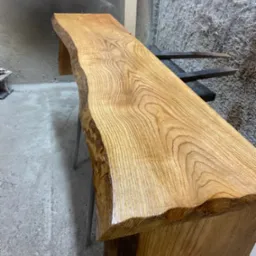 Surowy, lakierowany blat z litego drewna o nieregularnych krawędziach, ustawiony na metalowych nogach w warsztacie.