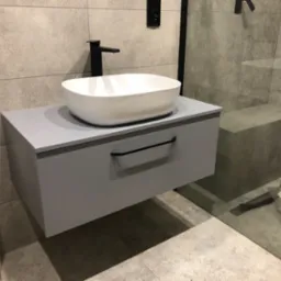 Nowoczesna łazienka: biała umywalka nablatowa na szarej szafce z czarnym uchwytem, czarna bateria i włącznik na tle szarych płytek imitujących kamień.