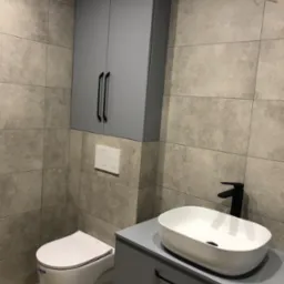 Nowoczesna łazienka z szarą zabudową, wiszącą szafką, umywalką nablatową i toaletą podwieszaną, wykończona płytkami imitującymi beton.
