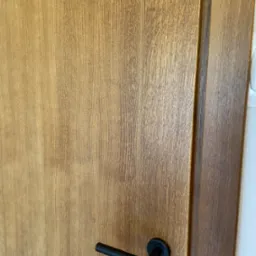 Drewniane drzwi wewnętrzne z czarną, minimalistyczną klamką, widoczna struktura drewna i ościeżnica.