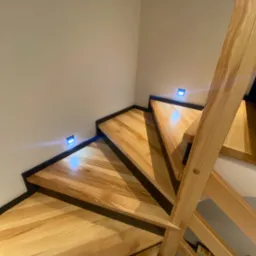 Nowoczesne schody drewniane z podświetleniem LED w ścianie, widok z g&oacute;ry, jasne drewno i czarne obramowanie stopni.