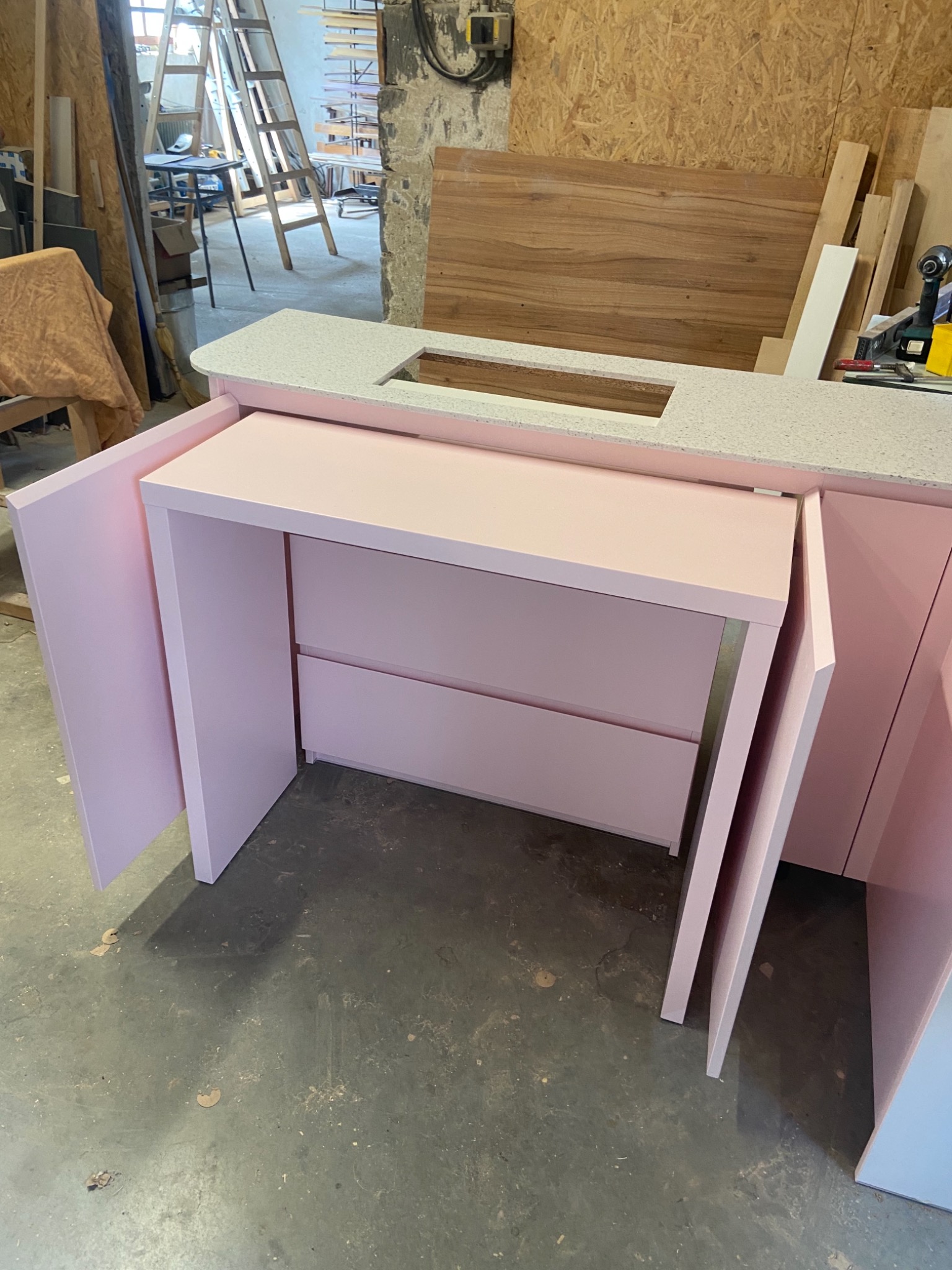 Różowa zabudowa meblowa z blatem w jasne plamki, widoczne otwarte drzwiczki szafek i szuflady, w tle warsztat stolarski z drabiną i narzędziami.